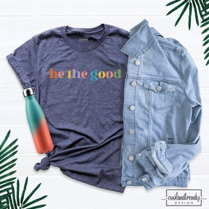 May include: A heather navy blue short-sleeve t-shirt with the text "be the good" in multicolored letters, paired with a light blue denim jacket and a teal and coral gradient metal water bottle with a silver cap.