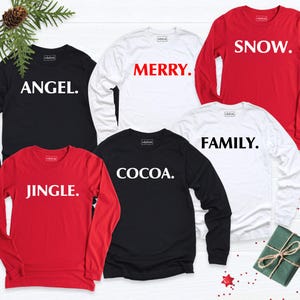 Pode incluir: Uma coleção de cinco camisas de manga comprida em vermelho, branco e preto. Cada camisa apresenta uma única palavra impressa numa fonte negrito sem serifa: ANGEL, MERRY, SNOW, JINGLE e COCOA, todas seguidas por um ponto.