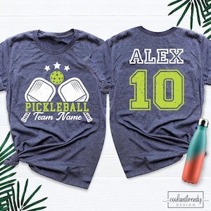 May include: A heather navy blue t-shirt featuring a front graphic of two pickleball paddles, a ball, three stars, and the text "PICKLEBALL Team Name". The back displays the name "ALEX" and the number "10" in large lime green font.