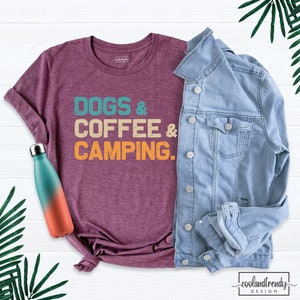 Pu&ograve; includere: Una maglietta color granata con il testo "Dogs & Coffee & Camping" in un font di stile vintage. Il testo &egrave; in tonalit&agrave; turchese, arancione e giallo. La maglietta &egrave; appoggiata su una superficie bianca con una giacca di jeans e una bottiglia d'acqua turchese e arancione.