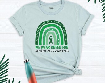 May include: A light blue t-shirt featuring a green rainbow graphic with a central awareness ribbon. Below the rainbow, the text reads WE WEAR GREEN FOR Cerebral Palsy Awareness. A gradient teal and orange metal water bottle is included.
