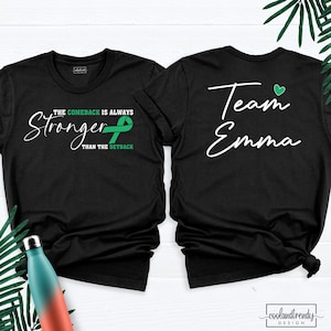 May include: Two black t-shirts. One shirt has the text "The Comeback is Always Stronger Than The Setback" with a green ribbon. The other shirt says "Team Emma" with a green heart. The shirts are on a white background.