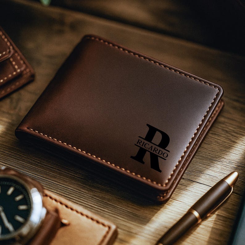 May include: A brown leather wallet with the name "Ricardo" and the letter "R" engraved on the front. The wallet is rectangular and has a stitched border. A watch and pen are also visible.