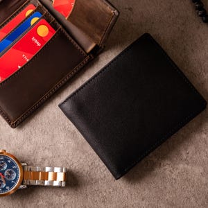May include: A black leather wallet with a stitched border is next to a brown leather wallet with visible credit cards. A gold and silver watch with a blue face is also in the image. The items are on a textured surface.