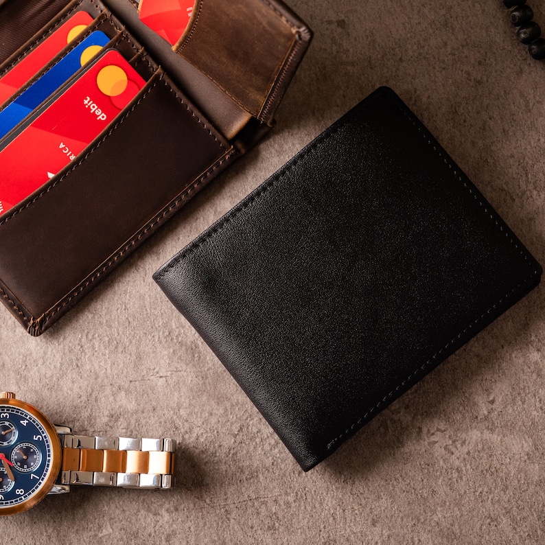 May include: A black leather wallet with a stitched border is next to a brown leather wallet with visible credit cards. A gold and silver watch with a blue face is also in the image. The items are on a textured surface.