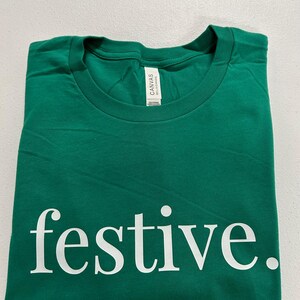 Puede incluir: Una camiseta verde doblada con la palabra "festive" impresa en blanco, utilizando una fuente sans-serif. La camiseta est&aacute; hecha de un material suave y tiene cuello redondo. La etiqueta dice "Canvas Bella+Canvas".