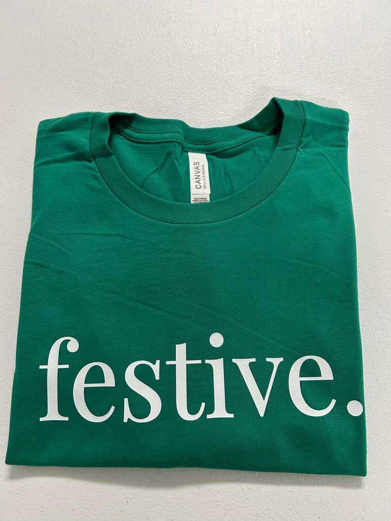 Puede incluir: Una camiseta verde doblada con la palabra "festive" impresa en blanco, utilizando una fuente sans-serif. La camiseta est&aacute; hecha de un material suave y tiene cuello redondo. La etiqueta dice "Canvas Bella+Canvas".