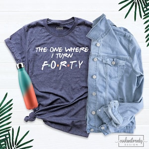 May include: A heather navy blue t-shirt with white text that reads THE ONE WHERE I TURN F O R T Y, accompanied by a light blue denim jacket and a gradient teal and coral metal water bottle with a silver cap.
