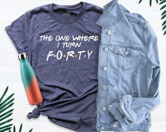 Pode incluir: Uma t-shirt azul-marinho mesclado com texto branco que diz THE ONE WHERE I TURN F O R T Y, acompanhada por um casaco de ganga azul claro e uma garrafa de água de metal com gradiente turquesa e coral e tampa prateada.