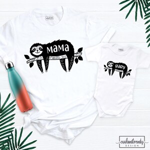 May include: White t-shirt with a black sloth design and the word "mama". A matching white baby onesie features a similar sloth design with the word "baby". A teal and orange water bottle is visible on the left.