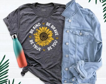 May include: A dark heather gray short-sleeve t-shirt featuring a large yellow sunflower graphic surrounded by small butterflies and the text BE KIND BE BRAVE BE TRUE BE YOU, paired with a light blue denim jacket and a teal and orange water bottle.