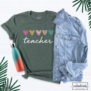 May include: A green t-shirt with the word "teacher" in white script, adorned with five colorful heart designs. A light blue denim jacket and a gradient water bottle are also in the image. The shirt is part of a teacher-themed collection.