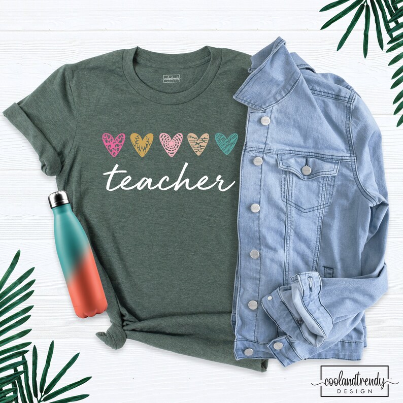 May include: A green t-shirt with the word "teacher" in white script, adorned with five colorful heart designs. A light blue denim jacket and a gradient water bottle are also in the image. The shirt is part of a teacher-themed collection.