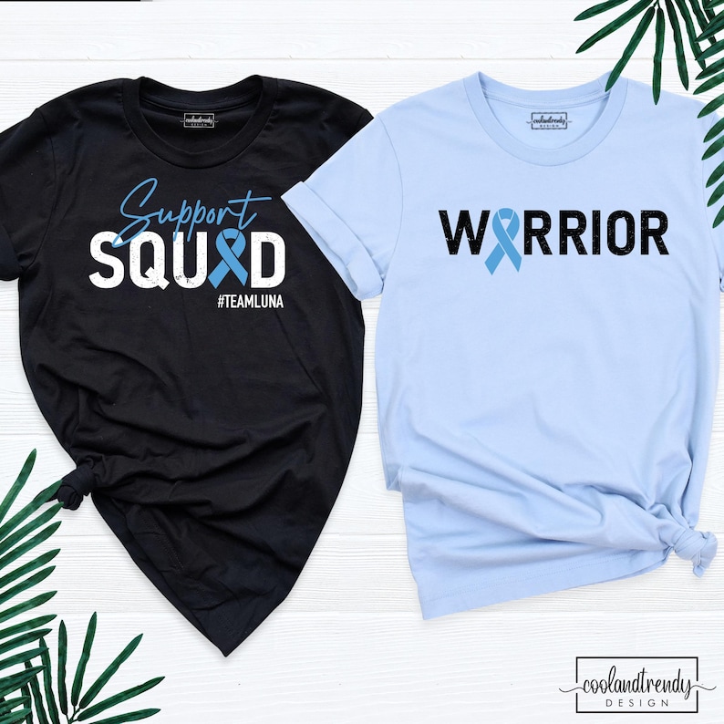 May include: Two t-shirts, one black and one light blue, with a blue ribbon design and text. The black shirt says "Support Squad #TeamLuna" and the light blue shirt says "Warrior".