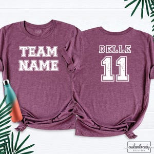 Puede incluir: Dos camisetas granates con letras blancas. Una camiseta dice "TEAM NAME" en la parte delantera. La otra camiseta tiene "BELLE" y el n&uacute;mero "11" en la espalda. Una botella de agua turquesa y naranja es visible a la izquierda.