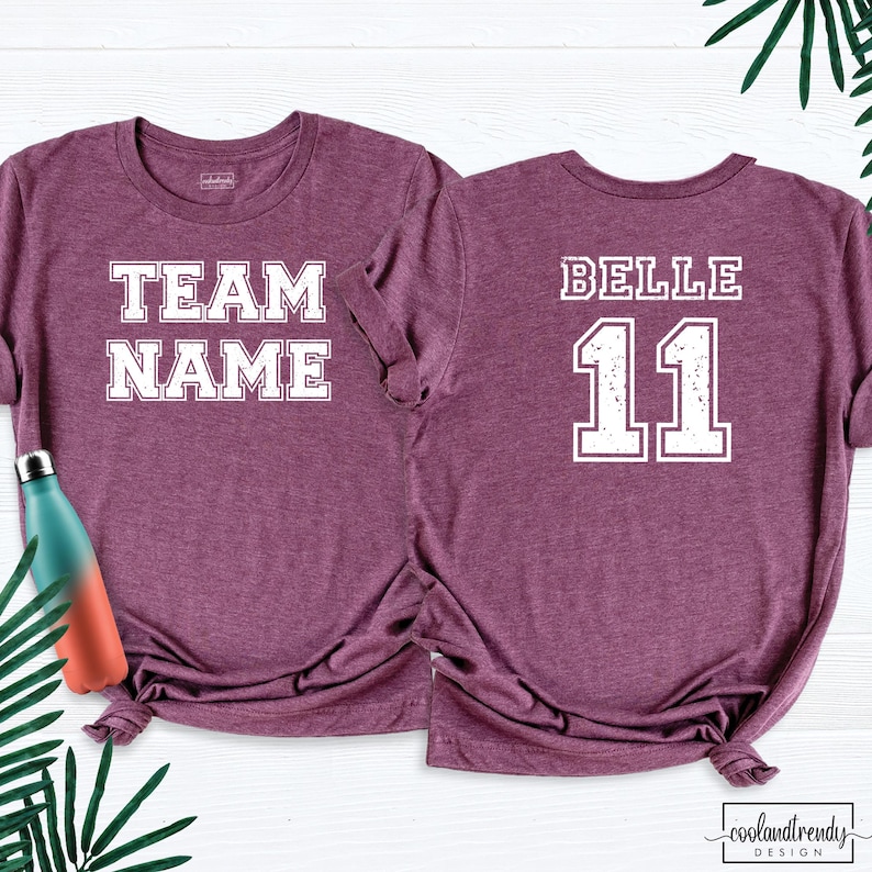 Puede incluir: Dos camisetas granates con letras blancas. Una camiseta dice "TEAM NAME" en la parte delantera. La otra camiseta tiene "BELLE" y el n&uacute;mero "11" en la espalda. Una botella de agua turquesa y naranja es visible a la izquierda.