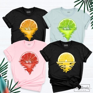Pode incluir: Quatro t-shirts de manga curta em preto, azul claro e rosa. Cada t-shirt apresenta um gráfico de uma fatia de citrino (laranja, lima, toranja ou limão) sobre um reflexo estilizado e escorrendo da fruta na cor correspondente.
