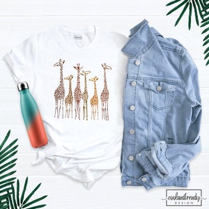 May include: White t-shirt featuring a graphic of a group of giraffes in a gradient of brown, orange, and yellow colours. The giraffes are standing in a line, with their heads turned to the side. The t-shirt is on a white surface, next to a blue denim jacket and a water bottle.