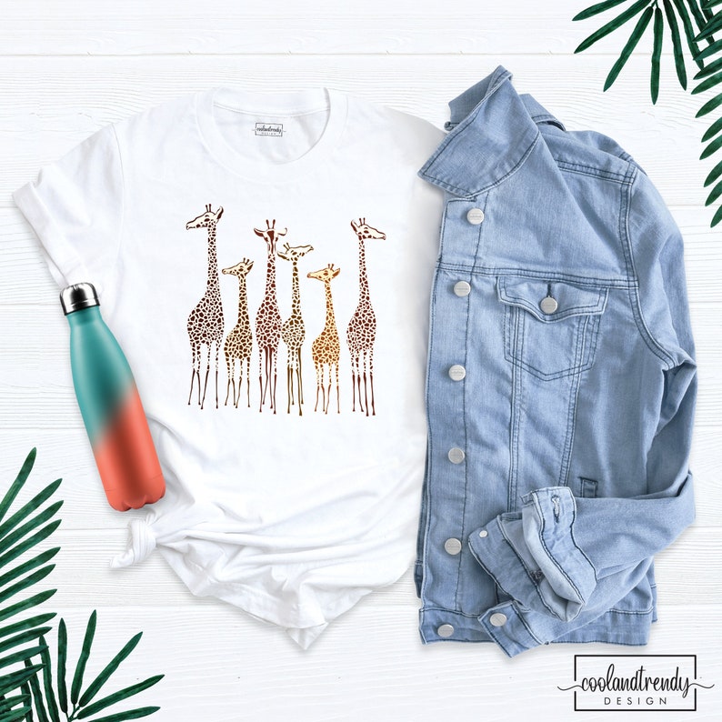 May include: White t-shirt featuring a graphic of a group of giraffes in a gradient of brown, orange, and yellow colours. The giraffes are standing in a line, with their heads turned to the side. The t-shirt is on a white surface, next to a blue denim jacket and a water bottle.