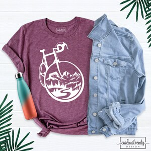 May include: A maroon t-shirt with a white graphic of a bicycle riding through a mountain landscape. The t-shirt is laid flat on a white wooden surface with a denim jacket and a water bottle.