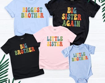 May include: A collection of five garments including four short-sleeve t-shirts and one baby bodysuit in light blue, black, and pink. The shirts feature colorful retro-style text reading BIGGEST BROTHER, BIG SISTER AGAIN, BIG BROTHER, LITTLE SISTER, and LITTLE BROTHER.