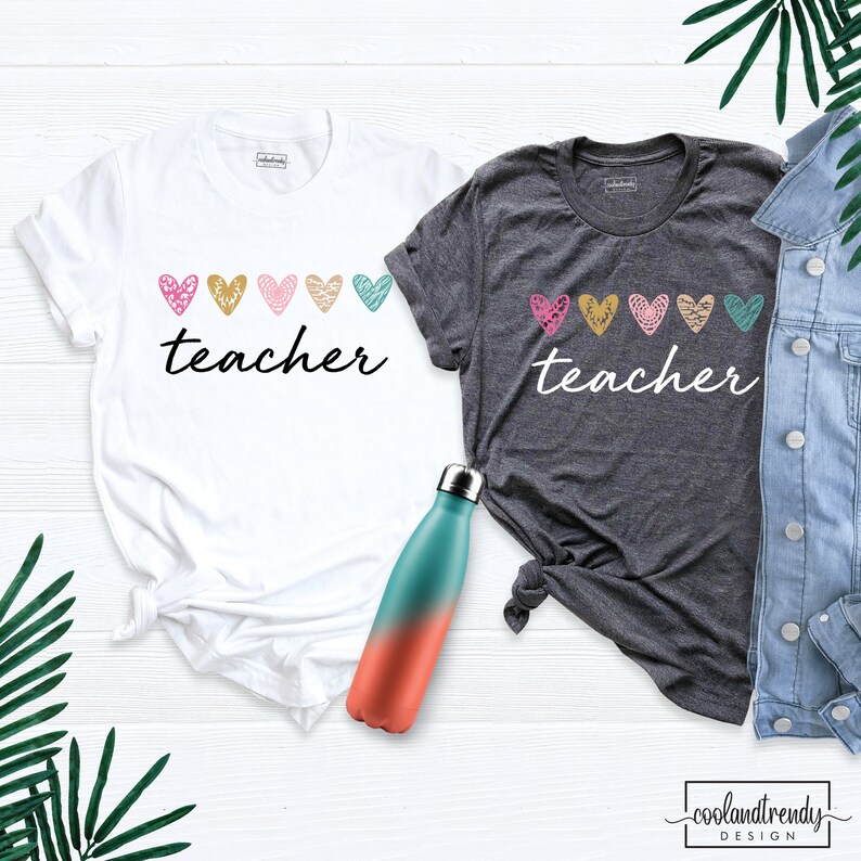 May include: Two t-shirts, one white and one charcoal gray, with the word "teacher" in a script font. Above the word are five colorful hearts. A teal and coral water bottle is in the foreground.