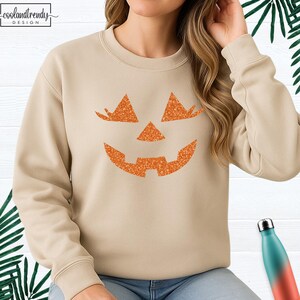 May include: Beige sweatshirt featuring a glittery orange jack-o'-lantern face design. The sweatshirt has a crew neck and long sleeves. The text "coolandtrendy DESIGN" is in the upper left corner. A seasonal apparel item for Halloween.