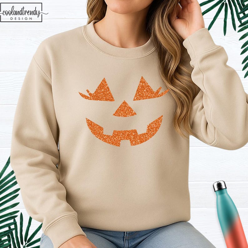 May include: Beige sweatshirt featuring a glittery orange jack-o'-lantern face design. The sweatshirt has a crew neck and long sleeves. The text "coolandtrendy DESIGN" is in the upper left corner. A seasonal apparel item for Halloween.