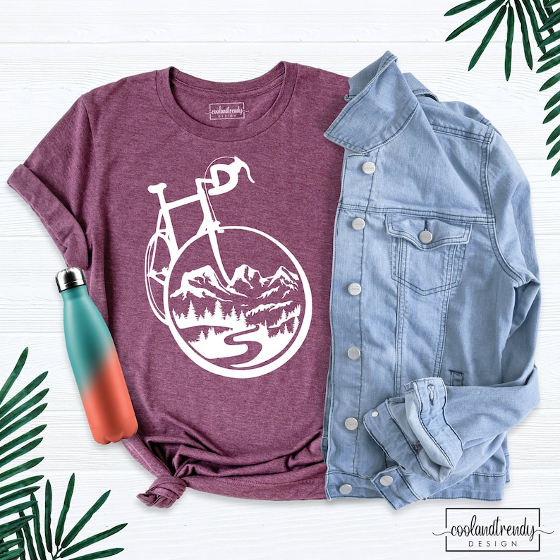 May include: A maroon t-shirt with a white graphic of a bicycle with mountains and a river in the background. The shirt is laid out on a white wooden surface with a blue denim jacket and a teal and orange water bottle.