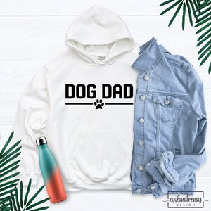 Puede incluir: Sudadera con capucha blanca con texto negro que dice "DOG DAD" con una huella de pata debajo. La sudadera est&aacute; sobre una superficie de madera blanca con una chaqueta vaquera y una botella de agua.