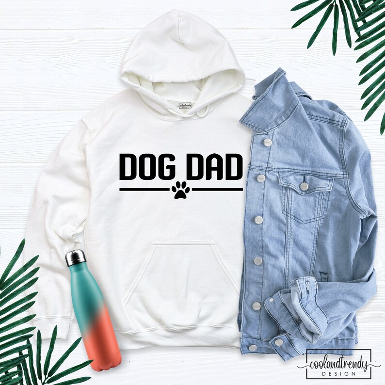 Puede incluir: Sudadera con capucha blanca con texto negro que dice "DOG DAD" con una huella de pata debajo. La sudadera est&aacute; sobre una superficie de madera blanca con una chaqueta vaquera y una botella de agua.