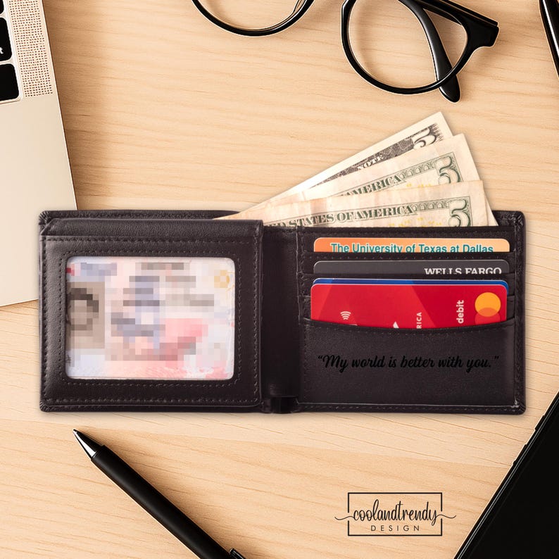 May include: Open black leather wallet with cash, credit cards, and a clear ID window. The wallet has the text "My world is better with you." printed on the inside. The wallet is on a wooden surface.