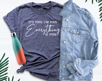 May include: A heather navy blue t-shirt with the white text IT'S FINE. I'M FINE. Everything IS FINE. A light blue denim jacket with silver buttons is layered over the shirt, and a teal and coral gradient metal water bottle is placed nearby.