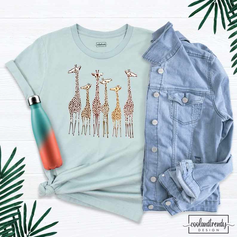 May include: A light blue t-shirt featuring a graphic of a group of giraffes in various shades of brown and orange. The giraffes are standing in a line, with their heads turned to the side. The t-shirt is a casual style with short sleeves and a crew neck.