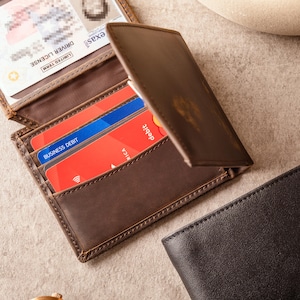 May include: A brown leather wallet, partially open, displaying a driver's license and credit cards. The wallet has multiple card slots. A second black leather wallet is visible in the background. The wallet is made of leather.