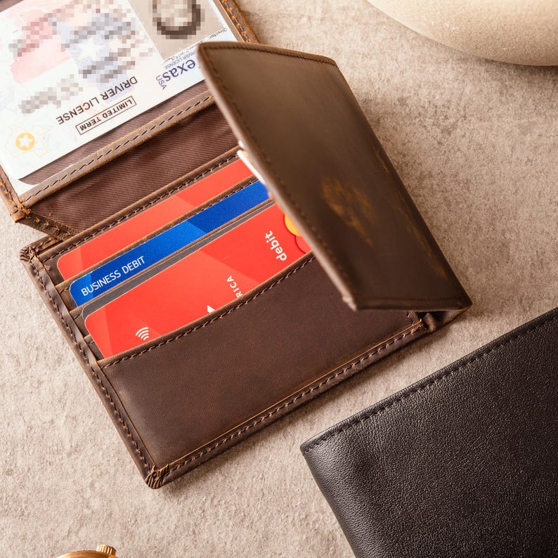 May include: A brown leather wallet, partially open, displaying a driver's license and credit cards. The wallet has multiple card slots. A second black leather wallet is visible in the background. The wallet is made of leather.