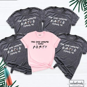 May include: Four dark gray t-shirts and one light pink t-shirt. The dark gray shirts feature the white text THE ONE WHERE K.A.T.I.E. TURNS 40. The light pink shirt features the black text THE ONE WHERE I TURN FORTY. Each letter has a small colored dot.