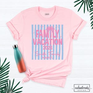 May include: A light pink t-shirt featuring a graphic with vertical light blue stripes, a palm tree silhouette, and the text "FAMILY VACATION 2026", "Smith Family", and "MIAMI FL". A teal and orange gradient water bottle is positioned to the left.