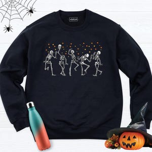 May include: A black crewneck sweatshirt featuring a graphic of five white skeletons dancing among small orange stars. The garment has long sleeves and a ribbed neckline. The interior label displays the text "enchanted threads DESIGN".