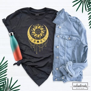 Puede incluir: Camiseta negra con un dise&ntilde;o de sol y luna de purpurina dorada. El sol tiene una cara y la luna tiene tres lunas crecientes colgando de ella.