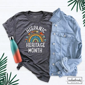 May include: A dark grey t-shirt with the text "HISPANIC HERITAGE MONTH" in white. A rainbow-colored graphic with flags of Hispanic countries is above the text.