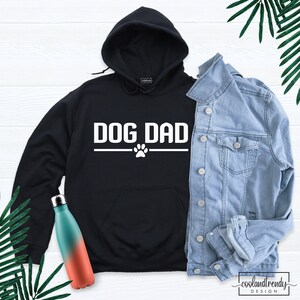 Puede incluir: Sudadera con capucha negra con el texto "DOG DAD" en blanco y una huella de pata debajo. La sudadera est&aacute; colocada sobre una superficie blanca con una chaqueta vaquera y dos botellas de agua.