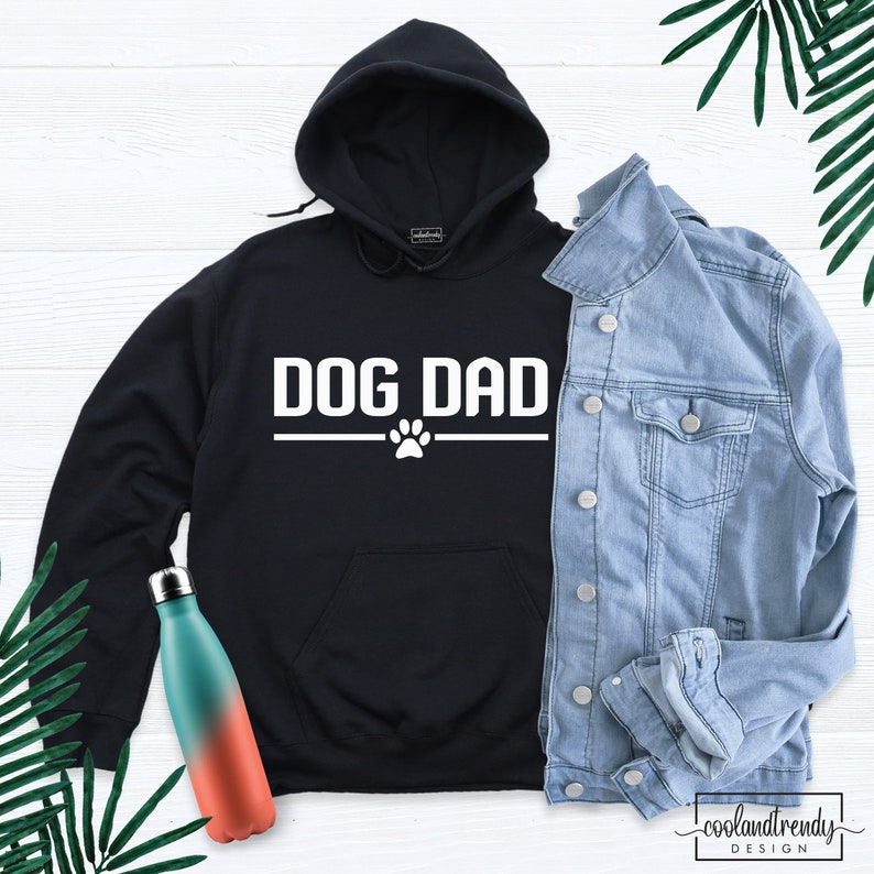 Puede incluir: Sudadera con capucha negra con el texto "DOG DAD" en blanco y una huella de pata debajo. La sudadera est&aacute; colocada sobre una superficie blanca con una chaqueta vaquera y dos botellas de agua.