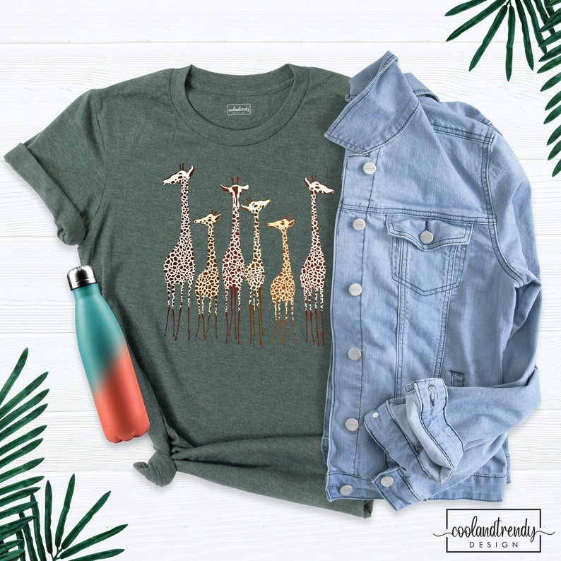 May include: A green t-shirt featuring a graphic print of a group of giraffes in various shades of brown and yellow. The giraffes are standing in a line, with their necks outstretched. The t-shirt is on a white wooden surface, next to a blue denim jacket and a teal and orange water bottle.