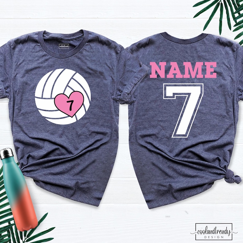May include: Two navy blue t-shirts. The front shirt has a volleyball graphic with a pink heart and the number 7. The back shirt has the word "NAME" and the number 7 in pink and white. The shirts are from coolandtrendy design.