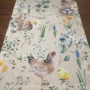 Puede incluir: Camino de mesa beige con temática de Pascua. El camino presenta ilustraciones de un conejo, gallinas, pollitos, huevos en un nido y varias flores y vegetación. El diseño incluye detalles florales amarillos, blancos y azules.