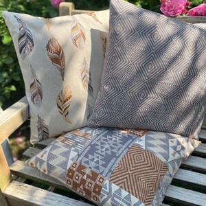 Puede incluir: Tres cojines cuadrados con patrones distintos. Uno tiene un fondo beige con ilustraciones de plumas marrones y negras. Otro es gris oscuro con un patr&oacute;n geom&eacute;trico de diamantes repetido. El tercero presenta un dise&ntilde;o de estilo patchwork.