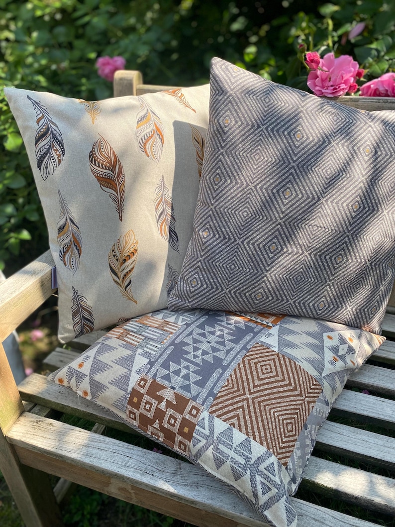 Puede incluir: Tres cojines cuadrados con patrones distintos. Uno tiene un fondo beige con ilustraciones de plumas marrones y negras. Otro es gris oscuro con un patr&oacute;n geom&eacute;trico de diamantes repetido. El tercero presenta un dise&ntilde;o de estilo patchwork.