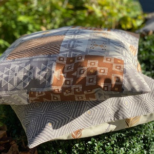 Puede incluir: Dos cojines cuadrados apilados uno sobre otro. El coj&iacute;n superior presenta un dise&ntilde;o de patchwork con patrones geom&eacute;tricos en marr&oacute;n, gris y blanco. El coj&iacute;n inferior tiene un patr&oacute;n de diamantes en gris y blanco con peque&ntilde;os detalles dorados.
