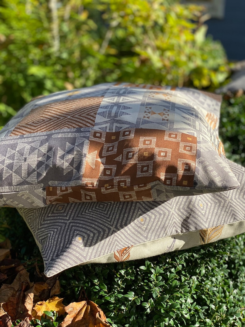Puede incluir: Dos cojines cuadrados apilados uno sobre otro. El coj&iacute;n superior presenta un dise&ntilde;o de patchwork con patrones geom&eacute;tricos en marr&oacute;n, gris y blanco. El coj&iacute;n inferior tiene un patr&oacute;n de diamantes en gris y blanco con peque&ntilde;os detalles dorados.